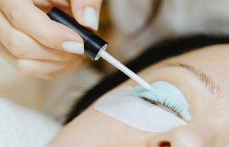 Girl cosmetologist laminates and paints woman eyelashes Portrait of a young unrecognizable Caucasian girl cosmetologist in a white coat who applies an adhesive solution to a silicone mold with a brush of the right eye of a brunette woman lying on a cosmetology table in a home beauty salon for eyelash lifting, side view close-up with selective focus.